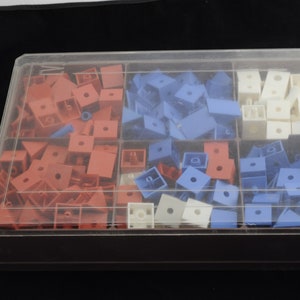 May include: A clear plastic box filled with colorful plastic blocks. The blocks are in four different colors: red, blue, white, and a light shade of blue. The box has four compartments, each filled with a different color of blocks.
