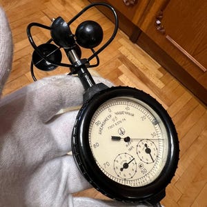 Può includere: Un anemometro nero e argento vintage con un indicatore circolare e tre quadranti più piccoli. L'indicatore legge "45" e include testo in cirillico. L'anemometro ha una serie di coppe nere e un manico in metallo.