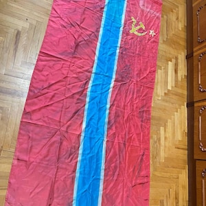 May include: A red flag with a central vertical blue stripe and white stripes either side. The flag has a yellow hammer and sickle symbol with a star in the upper right corner. The flag is on a wooden floor.
