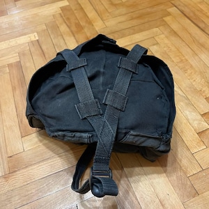 Puede incluir: Una mochila negra con correas ajustables y cierre de hebilla. La mochila está hecha de un material duradero, posiblemente lona. Las correas son de color verde oscuro y se cruzan en la espalda.