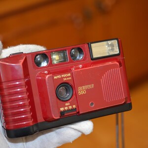 May include: A red and black 35mm film camera with a built-in flash. The camera has a lens with a 34mm focal length and says "Retue 550" on the front.