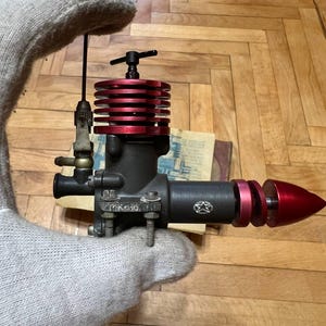 May include: A model airplane engine with a black body and red cooling fins. The engine has a silver-colored carburetor and a red cone-shaped nose. The engine is held by a gloved hand.
