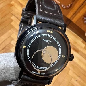 Puede incluir: Reloj de pulsera negro con correa de cuero. La esfera del reloj muestra la palabra "RAKETA" en blanco, un diseño de fase lunar dorado y detalles dorados. El reloj tiene un bisel negro y una corona negra.