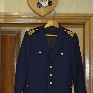 Könnte beinhalten: Ein dunkelblauer Blazer mit goldenen Knöpfen und goldenen Besätzen an den Schultern. Der Blazer hängt an einem Kleiderbügel vor einer Holzwand.
