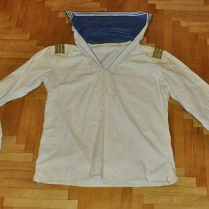 May include: A white long-sleeved shirt with a blue and white sailor collar and gold trim on the shoulders. The shirt has a V-neck and is made of a lightweight fabric.