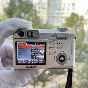 Può includere: Fotocamera digitale argentata con cinturino nero e giallo. Lo schermo della fotocamera mostra le opzioni del menu, tra cui dimensioni, qualità, nitidezza, contrasto e colore. Il marchio "TRAVELER" è visibile.