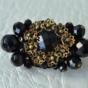May include: A black and gold brooch featuring a large black crystal at its centre, surrounded by smaller black beads and gold filigree.