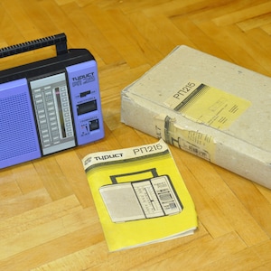 Puede incluir: Una radio de transistores morada y negra con asa, un manual de instrucciones amarillo y una caja de cartón marrón etiquetada "PT215".