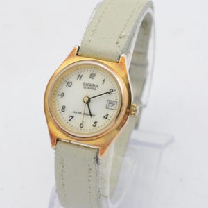 Puede incluir: Un reloj de pulsera analógico de tono dorado con una esfera blanca y una correa de cuero blanca. La esfera del reloj tiene agujas negras y marcadores de hora romanos negros. La esfera del reloj también tiene una ventana de fecha en la posición de las 3 en punto. La esfera del reloj dice "SHARP QUARTZ" y "WATER RESISTANT".