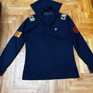 Könnte beinhalten: Ein dunkelblaues Langarm-Pullover-Shirt mit einem großen Matrosenkragen, goldenen Schulterklappen mit den Buchstaben CO, drei goldenen Streifen am rechten Ärmel und einem runden roten Aufnäher mit schwarzem Emblem am linken Ärmel.
