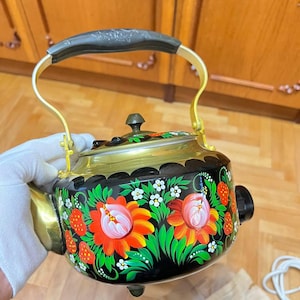 Puede incluir: Una tetera eléctrica negra con un diseño floral con flores rojas, naranjas y blancas, y hojas verdes. La tetera tiene un borde y soportes de asa dorados, con un asa negra. La tetera está decorada con un estilo de arte popular.