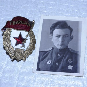 Può includere: Fotografia in bianco e nero di un uomo in uniforme militare, accanto a un distintivo della Guardia Sovietica. Il distintivo presenta una stella rossa, uno stendardo rosso con testo cirillico e una corona d'alloro.