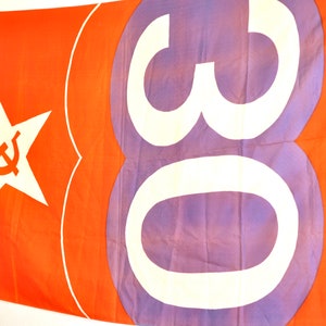 May include: A large orange flag with a white star and hammer and sickle symbol on the left side. The right side of the flag has a large white number "30" on a purple background.