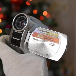Puede incluir: Una videocámara plateada con una lente negra y una etiqueta de DVD plateada y naranja que dice "30x Zoom Óptico".