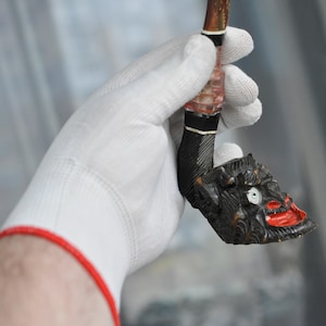 May include: A wooden pipe with a carved demon face and red painted lips. The pipe is held in a white glove with a red cuff.