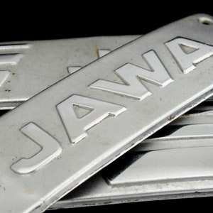 Puede incluir: Primer plano de una placa de metal plateado con la palabra "JAWA" en relieve. La placa está ligeramente curvada y tiene un aspecto desgastado y envejecido.