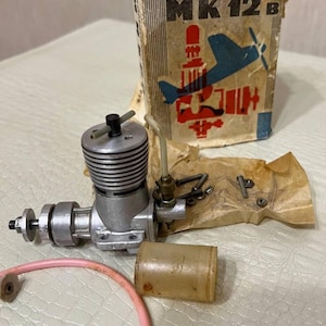 May include: A vintage model airplane engine with a silver finish, accompanied by a cardboard box with the text "MK 12 B" and an illustration of a blue airplane. A pink tube and a small fuel tank are also present.