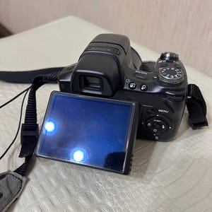 May include: A black digital camera with a lens, display screen, and strap. The screen is blue and reflects light. The camera has buttons and a dial on the top. The strap is black with a gray pad.