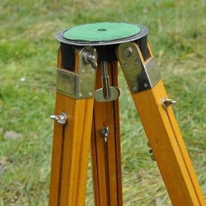 May include: A wooden tripod with a green felt top and metal accents. The tripod is made of wood and has a metal centre post.