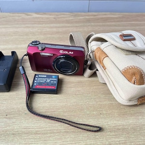 Könnte beinhalten: Eine rote Casio Exilim Digitalkamera mit 12,5-fach Zoom, ein schwarzes Batterieladegerät, ein Akku und eine beige Kameratasche mit einem braunen Lederaufnäher. Die Kamera und das Zubehör liegen auf einer Holzoberfläche.