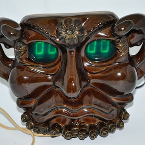 May include: A brown ceramic wall clock shaped like a grotesque face with green eyes that display the time with digital numbers. The clock has horns and a beard made of small brown ceramic balls.