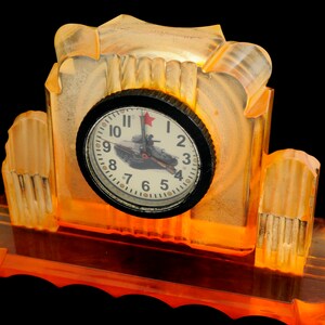 Puede incluir: Un pequeño reloj de color ámbar con una esfera negra y una estrella roja. La esfera del reloj presenta una imagen en blanco y negro de un tanque con una estrella roja en la parte superior. El reloj está sobre una base con un diseño decorativo.