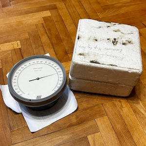 May include: A round analog gauge with a white face, black needle, and numerical markings from 80 to 105, resting on a white paper document. Beside it is a rectangular white styrofoam block with handwritten text on the top surface.