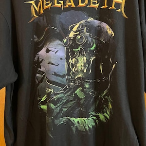 May include: Black t-shirt featuring the band name "MEGADETH" in gold lettering above a graphic. The graphic depicts a skull wearing a helmet with green-tinted goggles, and a soldier in green and black.