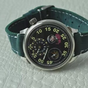 May include: A silver-cased wristwatch with a black face and a green leather strap. The watch face features multiple sub-dials and white numerals. The strap is dark green with white stitching. The watch has a classic, vintage aesthetic.