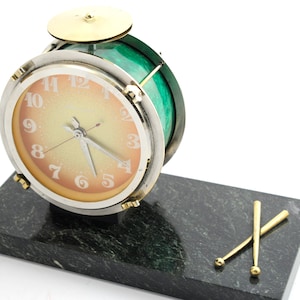 Può includere: Un orologio da tavolo verde e argento a forma di tamburo con un piatto di ottone in cima. L'orologio è su una base di marmo nero con due bacchette dorate.