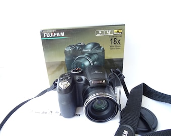 May include: Black Fujifilm digital camera with a lens cap and strap. The camera is in front of its box, which displays the camera's image and features, including 18x optical zoom and a 3.0-inch screen.
