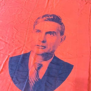May include: A red flag with a portrait of a man in a dark blue suit and tie. The portrait is a stylized image with a blue and white colour scheme, set against the solid red background of the flag.