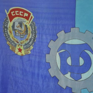 May include: A blue and turquoise flag with a large red, yellow, and white emblem featuring the text "CCCP" and the words "ПРОЛЕТАРИИ ВСЕХ СТРАН, СОЕДИНЯЙТЕСЬ!"  The emblem also includes a hammer and sickle.  A large gray gear with a blue and turquoise symbol is on the right side of the flag.