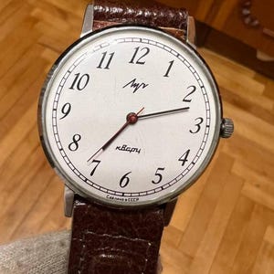 Puede incluir: Un reloj de pulsera vintage con esfera blanca, números negros y correa de cuero marrón. La esfera del reloj presenta el nombre de la marca y la palabra "Kvarz" en cirílico. El reloj tiene una caja plateada y un segundero rojo.
