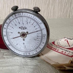 Puede incluir: Una regla de cálculo circular plateada vintage con marcas negras y la palabra "MATИKA" en la esfera. Se ve una funda roja y un papel con texto ruso. La regla de cálculo tiene dos perillas en la parte superior.