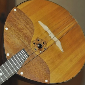 May include: A wooden balalaika, a Russian stringed instrument, featuring a triangular body and a long neck with frets. The instrument has a light brown finish with darker wood accents and six strings. White dots mark the frets.