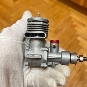 May include: A small, silver-colored model engine with a red cap and a black bolt. The engine has a ribbed cylinder head and a series of vents. It is held in a white glove.