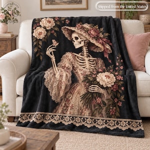 May include: A black plush throw blanket featuring a central graphic of a skeleton wearing a wide-brimmed floral hat and a lace-trimmed dress. The skeleton holds a bouquet of flowers and a butterfly. The blanket has a decorative lace border.