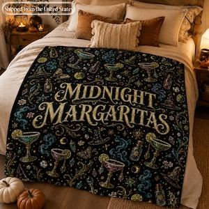 Op de afbeelding: Een zwarte plaid met de tekst Midnight Margaritas in gouden letters, omringd door een herhalend patroon van margaritaglazen, heksenhoeden, kristallen, vlinders en wervelende rook in tinten groen, paars en goud.