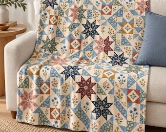May include: A quilt featuring a repeating pattern of eight-pointed stars and floral motifs in shades of navy blue, dusty rose, sage green, and mustard yellow on a cream background, draped over a light-colored sofa with a blue pillow.