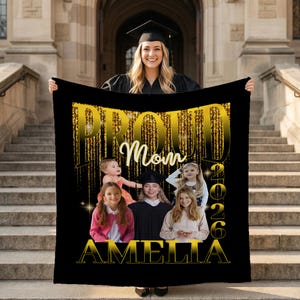 May include: A black blanket with gold text that reads "PROUD Mom" and "AMELIA 2026". The blanket features photos of children. A woman in a graduation cap holds the blanket in front of a stone building.