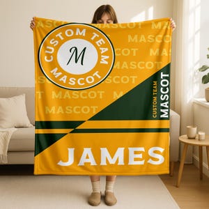 Könnte beinhalten: Eine gelb-grüne Decke mit dem Text "CUSTOM TEAM MASCOT" in einem Kreis und dem Namen "JAMES" in großen weißen Buchstaben. Die Decke hat einen diagonalen grünen Streifen und horizontale grüne Streifen.