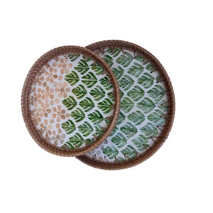 Puede incluir: Dos bandejas redondas decorativas con bordes marrones tejidos. Una bandeja presenta un patrón de pequeñas flores beige, mientras que la otra muestra un diseño de hojas verdes sobre fondo blanco. Las bandejas se superponen.