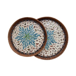 Puede incluir: Dos posavasos redondos decorativos. Cada posavasos presenta un diseño de mosaico con un patrón floral azul central y un borde tejido marrón. Los posavasos están sobre un fondo blanco.