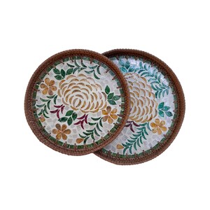 Puede incluir: Dos posavasos redondos con un diseño floral de mosaico. Los posavasos tienen un borde tejido marrón y un patrón central de mosaico de flores y hojas en tonos beige, verde y rojo. El fondo es blanco.