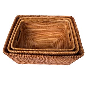 May include: Three rectangular woven rattan baskets of varying sizes, nested together. The baskets are a warm, natural brown color and feature built-in handles. The weave is tight and detailed, creating a textured surface.