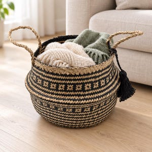 Könnte beinhalten: Ein runder Korb aus geflochtenem Seegras mit einem geometrischen Muster in Schwarz und Naturbeige. Der Korb hat zwei gedrehte Griffe und eine schwarze Quaste an einer Seite. Darin liegen eine cremefarbene Strickdecke und eine grüne Decke.