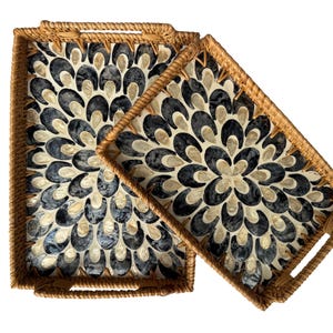 May include: Two rectangular serving trays with woven rattan frames. The tray interiors feature a floral pattern made of black, cream, and gray shell pieces. The trays are different sizes and are placed at an angle.