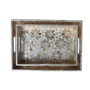 Pode incluir: Duas bandejas decorativas retangulares aninhadas. As bandejas têm um design floral em mosaico em tons de bege e branco. As bordas são de cor castanho claro com alças recortadas.