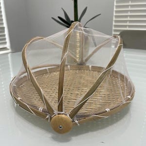May include: A round, woven bamboo food cover with a fine mesh net. The cover has a wooden handle and a hinged design for easy access. The bamboo has a natural, light brown colour, and the net is white.
